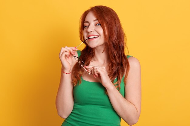 Mujer sonriente coqueteando mordiendo el marco de sus gafas y, vistiendo ropa casual