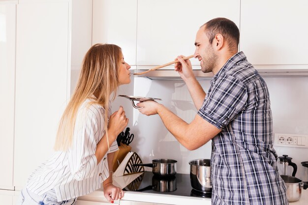 Mujer sonriente dejando que el hombre pruebe una sopa con una cuchara de madera en la cocina