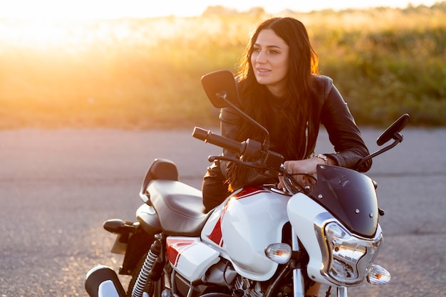 Mujer sonriente descansando sobre su motocicleta en la puesta de sol