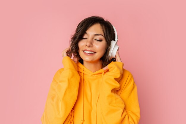 Mujer sonriente dichosa en auriculares blancos escuchando música en rosa. El uso de sudadera con capucha naranja.