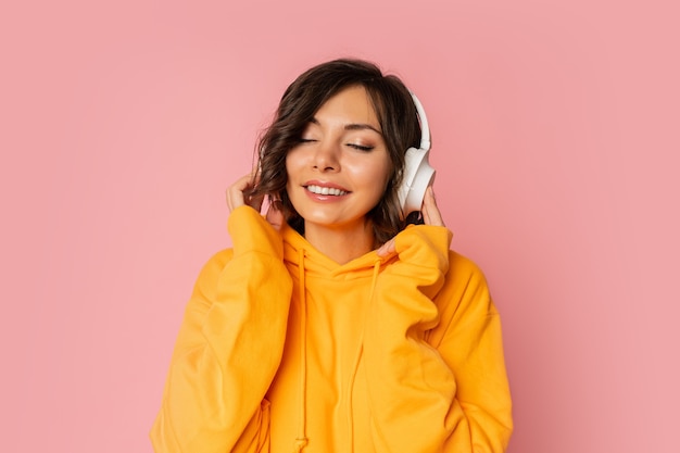 Mujer sonriente dichosa en auriculares blancos escuchando música en rosa. El uso de sudadera con capucha naranja.