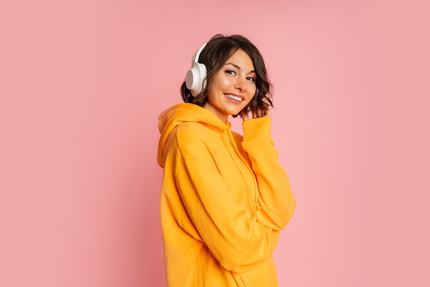 Mujer sonriente dichosa en auriculares blancos escuchando música en rosa. El uso de sudadera con capucha naranja.