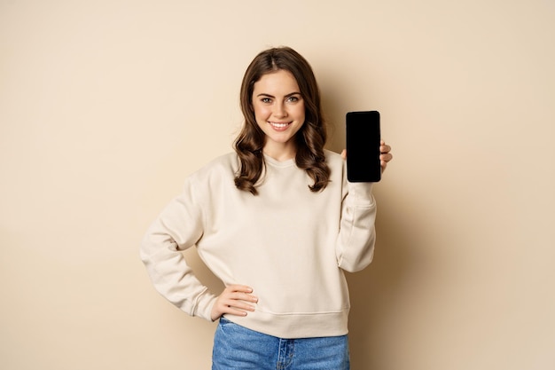 Mujer sonriente elegante que muestra la interfaz de la aplicación móvil de la pantalla del teléfono inteligente de pie sobre fondo beige