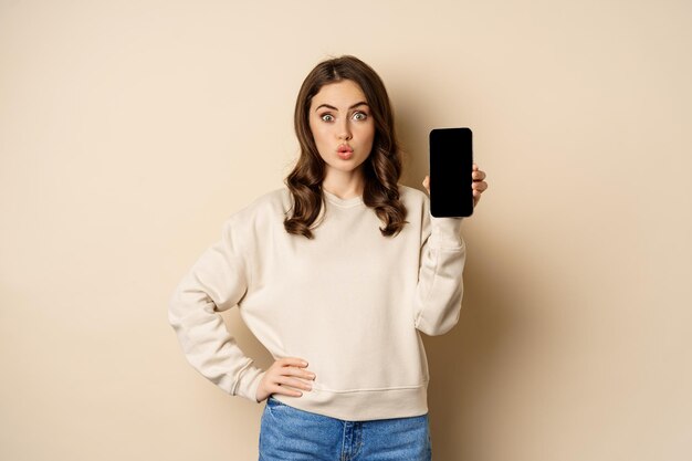 Mujer sonriente elegante que muestra la interfaz de la aplicación móvil de la pantalla del teléfono inteligente de pie sobre fondo beige