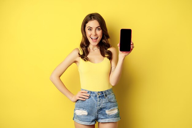 Mujer sonriente elegante que muestra la pantalla del teléfono móvil, la interfaz de la aplicación en el teléfono inteligente, de pie en la parte superior del tanque y pantalones cortos contra el fondo amarillo.