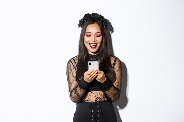 Mujer sonriente emocionada mirando el teléfono móvil complacido, con un vestido de encaje gótico para la fiesta de halloween, de pie sobre fondo blanco.