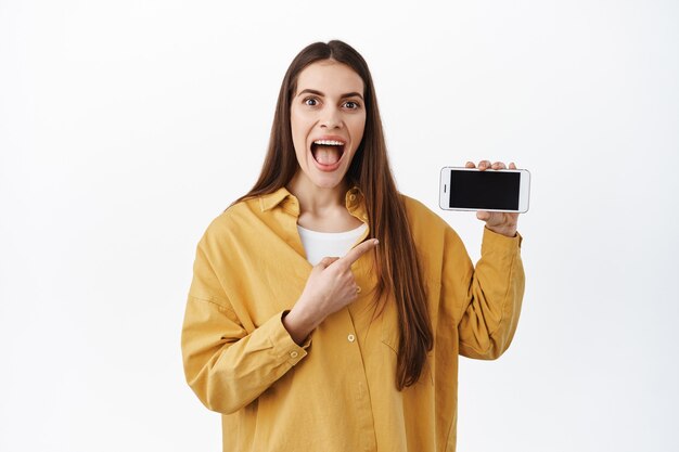 Mujer sonriente emocionada que muestra una aplicación impresionante, una aplicación de compras en línea, que señala con el dedo a la pantalla horizontal del teléfono inteligente, muestra algo sorprendente en la pantalla del teléfono, pared blanca