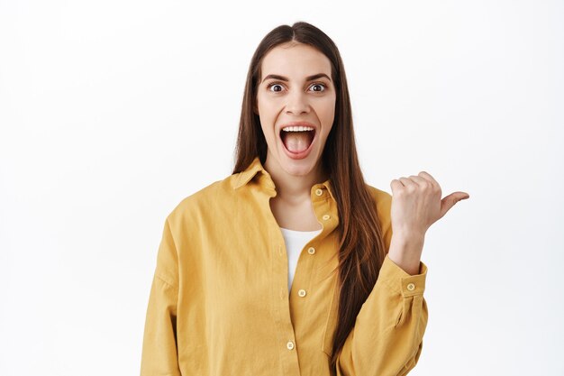 Mujer sonriente emocionada que muestra una oferta increíble, la mejor oferta en el mercado, apuntando con el pulgar a un lado en el espacio de la copia derecha, publicidad, de pie sobre una pared blanca