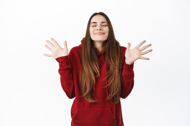 Mujer sonriente encantada cierra los ojos y siente satisfacción, levanta las manos y respira aire libremente, de pie feliz y despreocupado, regocijándose, de pie sobre una pared blanca