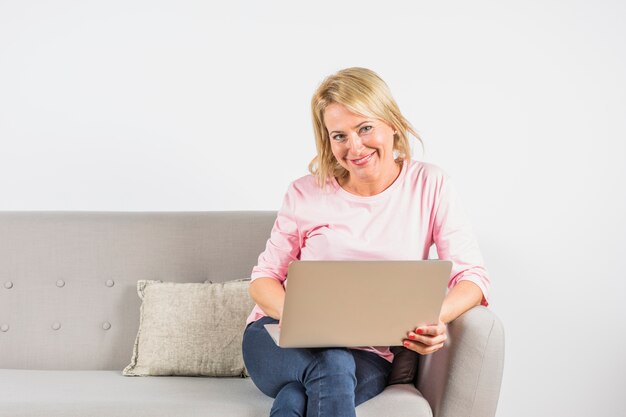 Mujer sonriente envejecida en blusa color de rosa con la computadora portátil en el sofá