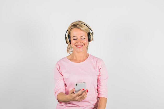 Mujer sonriente envejecida en blusa rosa con auriculares usando teléfono inteligente