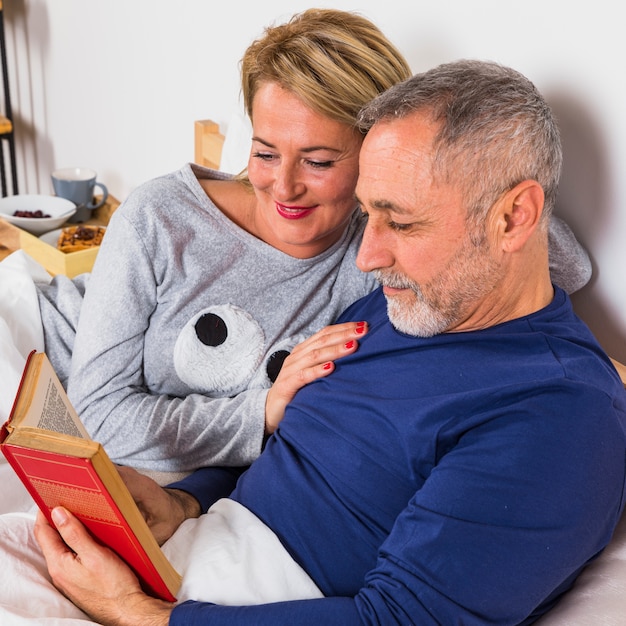 Mujer sonriente envejecida cerca del hombre con el libro en el edredón en cama