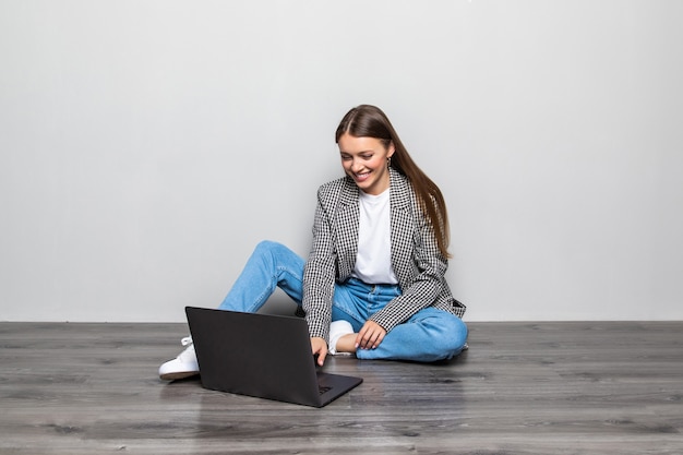 Mujer sonriente escribiendo en la computadora portátil mientras está sentado en el suelo con las piernas cruzadas aislado