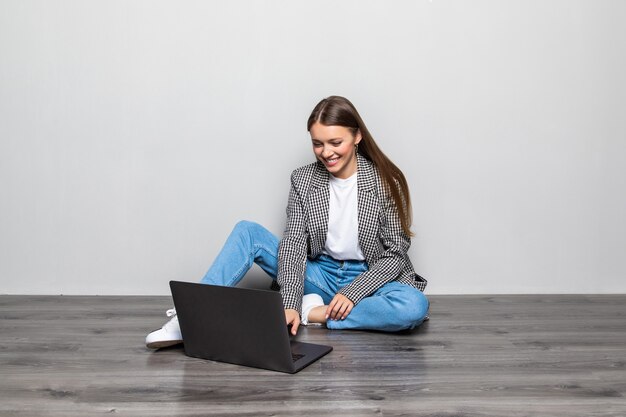 Mujer sonriente escribiendo en la computadora portátil mientras está sentado en el suelo con las piernas cruzadas aislado