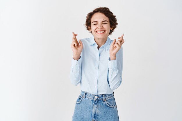 La mujer sonriente esperanzada cierra los ojos y cruza los dedos para la buena suerte, pidiendo deseos o soñando optimista, de pie en la pared blanca