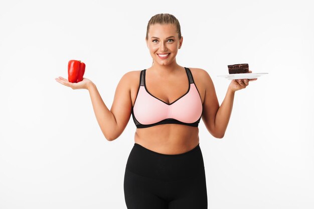 Mujer sonriente con exceso de peso en la parte superior deportiva con pimiento rojo y pastel de chocolate en las manos mientras mira felizmente en cámara sobre fondo blanco.