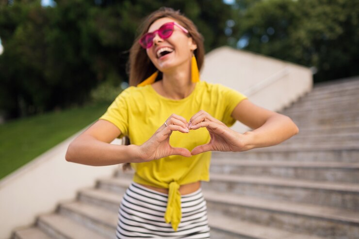 Mujer sonriente feliz bastante elegante joven que se divierte en el parque de la ciudad, positiva, emocional, con top amarillo, minifalda a rayas, gafas de sol rosas, tendencia de moda de estilo veraniego, mostrando el signo del corazón