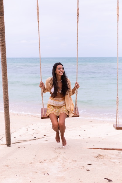 Mujer sonriente feliz en columpio en la playa, luz cálida del día.