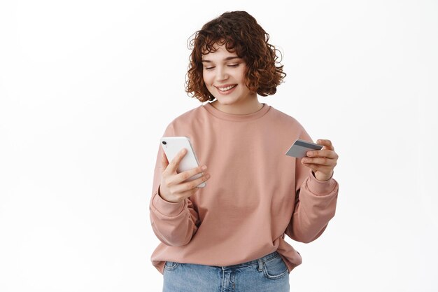 Mujer sonriente feliz mirando el teléfono inteligente, pagando compras en línea, con tarjeta de crédito, paga en la tienda de Internet desde la aplicación de teléfono móvil, de pie sobre fondo blanco.