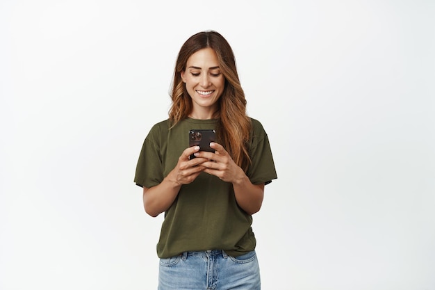 Foto gratuita mujer sonriente feliz, mujer adulta mirando su teléfono móvil, chateando en la aplicación, enviando mensajes, usando la aplicación de compras en línea, de pie contra un fondo blanco.