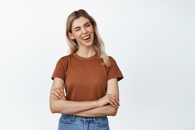 Mujer sonriente feliz con el pelo rubio guiñando un ojo y mirando con entusiasmo a la cámara de pie en camiseta marrón y jeans contra fondo blanco.