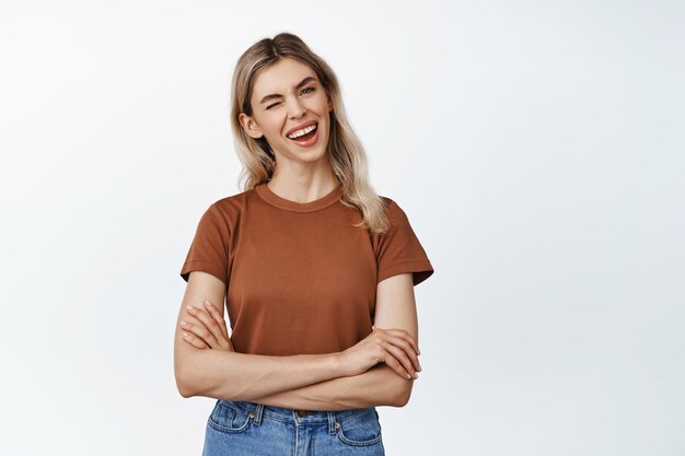 Mujer sonriente feliz con el pelo rubio guiñando un ojo y mirando con entusiasmo a la cámara de pie en camiseta marrón y jeans contra fondo blanco.