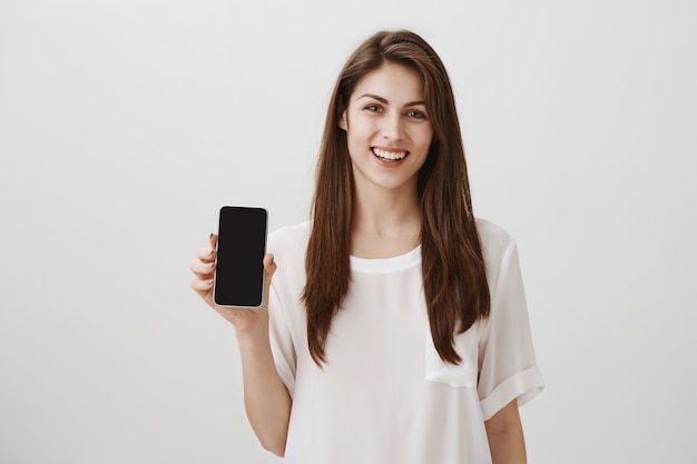 Mujer sonriente feliz que muestra la pantalla del móvil, recomienda la aplicación o el sitio de compras