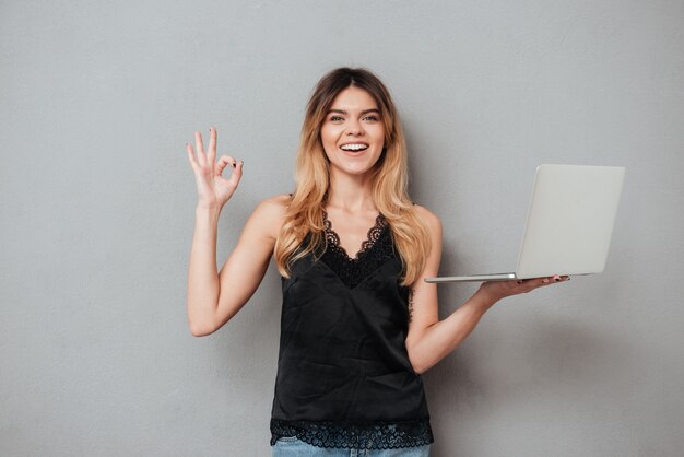 Mujer sonriente feliz que sostiene la computadora portátil y que muestra gesto aceptable