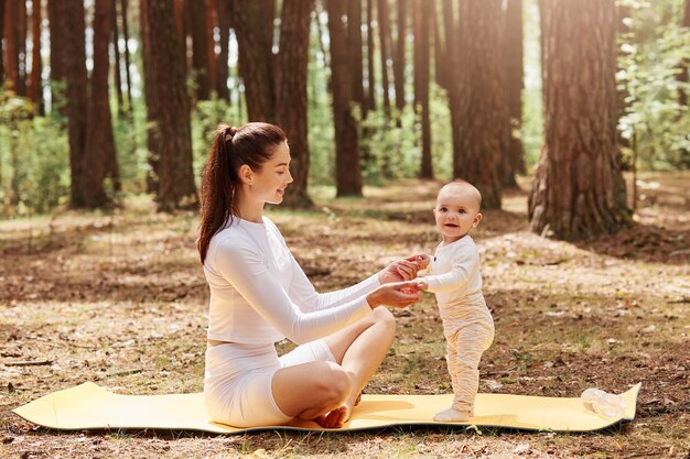 Mujer sonriente feliz en ropa deportiva de moda blanca sentada en la estera del gimnasio al aire libre, sosteniendo las palmas de los niños