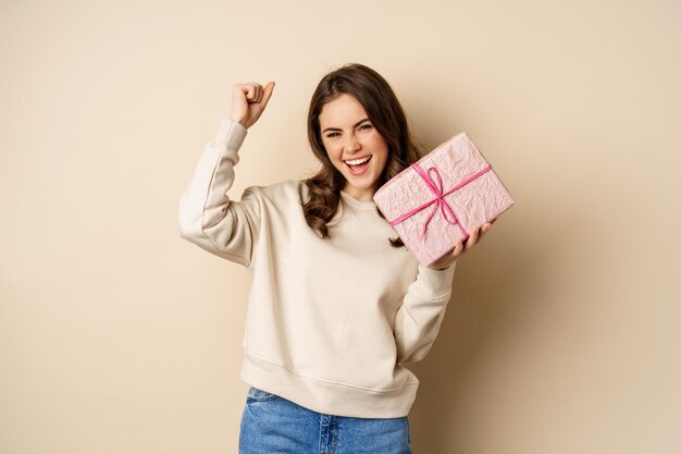 Mujer sonriente feliz sosteniendo un regalo y triunfando, levantando la mano alegremente, recibe un regalo de vacaciones, de pie sobre un fondo beige.