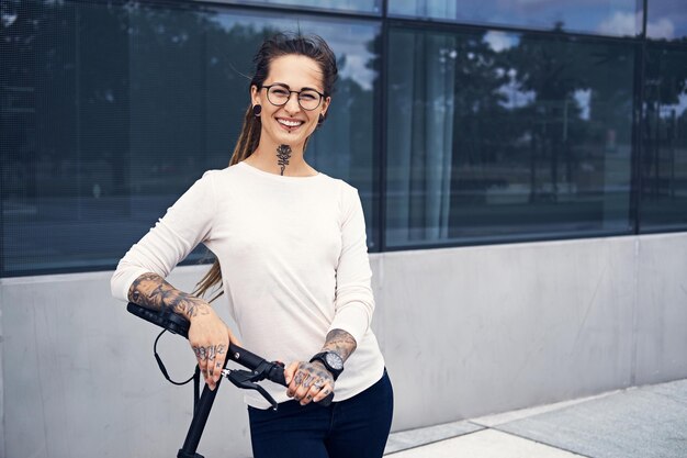 Mujer sonriente feliz con tatuajes y rastas conduce scooter eléctrico en la calle.