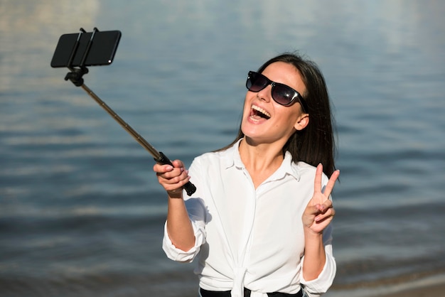 Mujer sonriente con gafas de sol tomando selfie en la playa y haciendo el signo de la paz