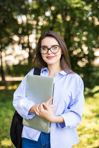 Foto gratuita mujer sonriente en gafas transparentes se queda con su computadora portátil en el parque