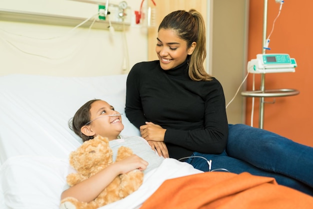 Mujer sonriente hablando con su hija enferma mientras se sienta en la cama en el hospital