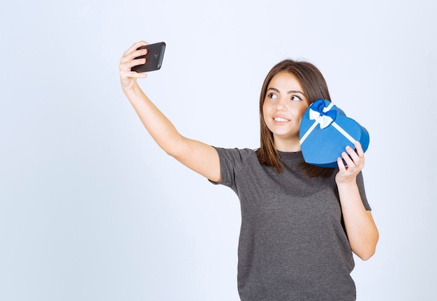 Una mujer sonriente haciendo selfie con una caja de regalo en forma de corazón.