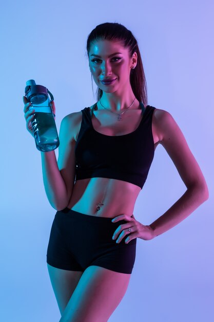 Mujer sonriente con hermoso cuerpo beber agua después del entrenamiento, aislado en luz púrpura con copyspace para texto