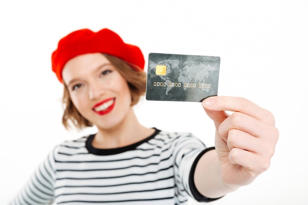 Mujer sonriente del jengibre que muestra la tarjeta de crédito en la cámara sobre gris. Centrarse en la tarjeta