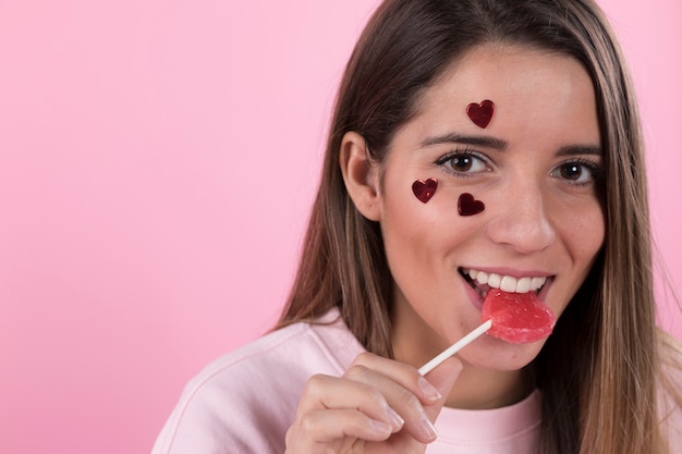 Mujer sonriente joven con los corazones del ornamento en cara y la piruleta