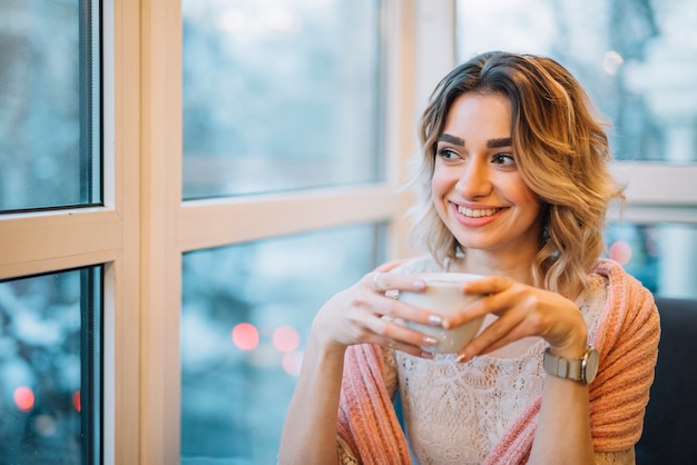 Foto gratuita mujer sonriente joven elegante con la taza de bebida cerca de la ventana en café