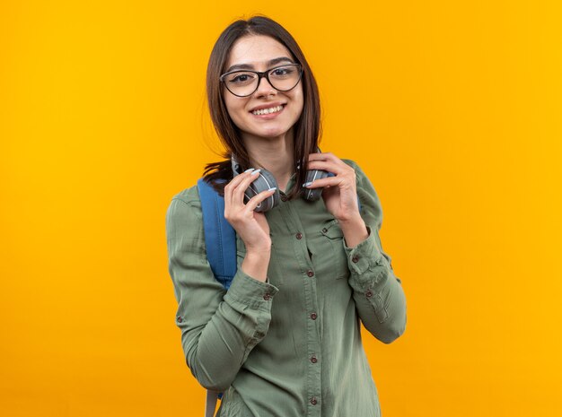 Mujer sonriente joven de la escuela con mochila con gafas y auriculares en el cuello