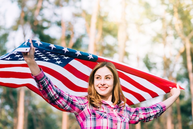 Mujer sonriente joven que lleva la bandera de los EEUU el Día de la Independencia