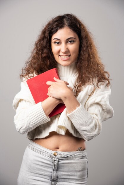 Mujer sonriente joven que sostiene el libro rojo sobre una pared gris. Foto de alta calidad