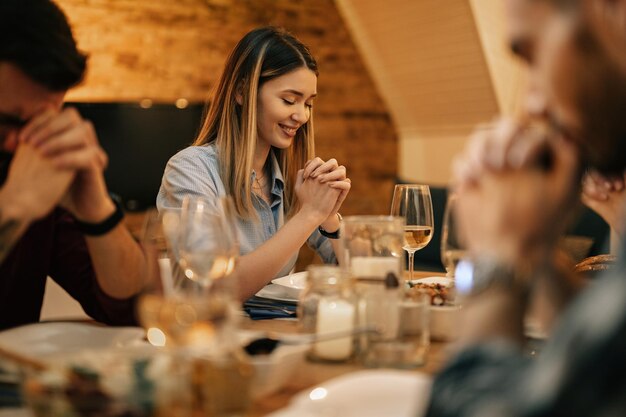 Mujer sonriente joven y sus amigos sentados en la mesa del comedor y rezando antes de una comida