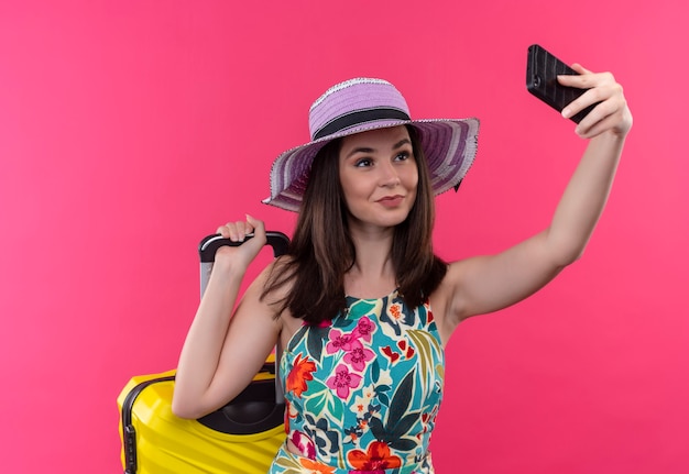 Mujer sonriente joven viajero haciendo selfie sosteniendo la maleta en la pared rosa aislada