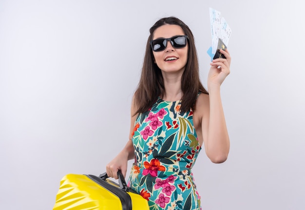 Mujer sonriente joven viajero en vestido multicolor con gafas sosteniendo una bolsa móvil y boletos en la pared blanca