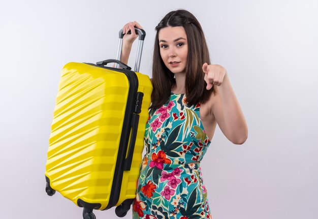 Mujer sonriente joven viajero con vestido multicolor sosteniendo una bolsa móvil y apunta hacia adelante en la pared blanca