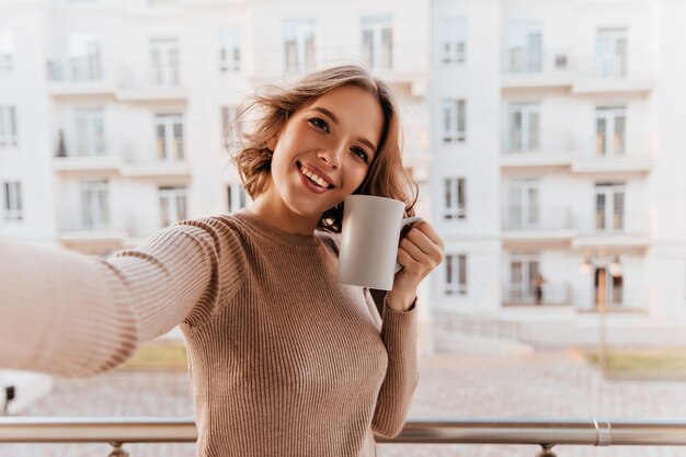 Mujer sonriente maravillosa con taza de café de pie en la ciudad. Chica morena positiva disfrutando de la mañana con té.