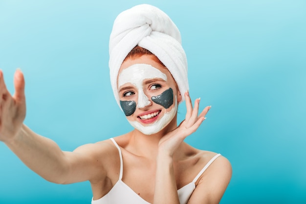 Mujer sonriente con mascarilla tomando selfie. Disparo de estudio de dama alegre con una toalla en la cabeza posando sobre fondo azul.