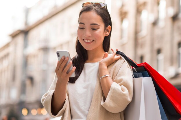 Mujer sonriente mirando smartphone al aire libre mientras sostiene bolsas de la compra.