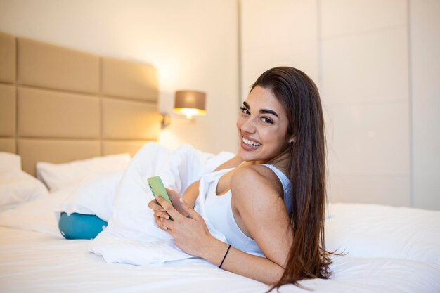 Mujer sonriente mirando el teléfono móvil mientras está acostada en la cama blanca Mujer joven morena feliz usando el teléfono celular en casa